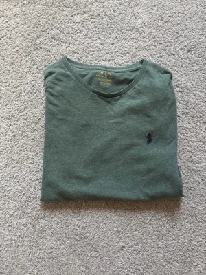 Grön t-shirt från Polo Ralph Lauren - Snygg grön t-shirt från Polo Ralph Lauren med klassisk rund hals och liten mörkblå logga broderad på bröstet. Tillverkad i mjuk bomull och har en normal passform. Perfekt basplagg för en avslappnad stil.