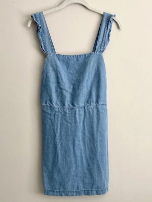 Ljusblå jeansjumpsuit med volang - Säljer en ljusblå jeansjumpsuit i mjukt denimtyg. Modellen har breda axelband med volangdetaljer och knappar framtill för justering. Klassisk rak passform och fickor på sidorna. Perfekt för en avslappnad och trendig look.