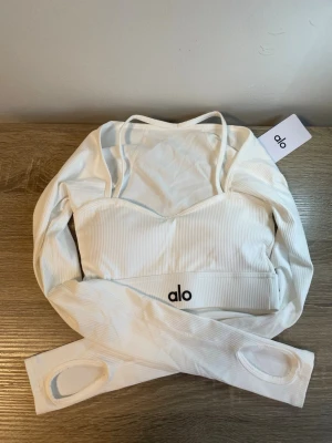 Vit ribbad långärmad topp från Alo - Snygg vit ribbad topp från Alo med långa ärmar och tumhål. Toppen har smala axelband och croppad passform, perfekt för en sportig och trendig look. Diskret Alo-logga framtill och stretchigt material som sitter bekvämt.