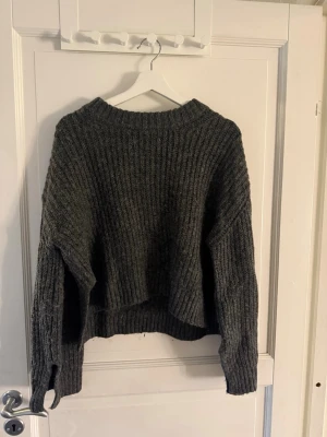 Mörkgrå stickad tröja  - Mysig mörkgrå stickad tröja med bred ribbstickning och avslappnad passform. Tröjan har rund halsringning, långa ärmar och något längre bakstycke. Perfekt för kalla dagar och enkel att styla till jeans eller kjol.