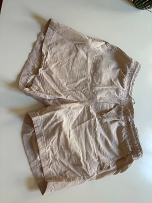 Beige linneshorts med resårmidja - Säljer ett par ljusbeige shorts i linne med avslappnad passform och elastisk midja med snörning. Shortsen har fickor på sidorna och en bakficka. Perfekta för varma sommardagar och riktigt sköna att bära.