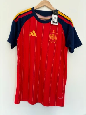 Spaniens matchtröja Adidas röd M - Säljer en officiell röd matchtröja från Spaniens landslag, tillverkad av Adidas. Tröjan har gula och blå detaljer, tunna vertikala ränder och landslagets emblem på bröstet. Tillverkad i lätt och ventilerande polyester, perfekt för fotboll eller träning.