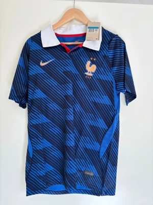 Frankrike Nike fotbollströja blå - Säljer en blå och svart randig fotbollströja från Nike för Frankrikes landslag. Tröjan har vit krage, röda detaljer och guldiga loggor, inklusive Nike Swoosh och Frankrikes tupp-emblem med två stjärnor. Tillverkad i Dri-FIT material för bästa komfort.