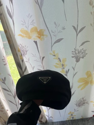 Svart keps från Prada - Säljer en svart keps från Prada med den klassiska trekantiga logotypen framtill. Kepsen har en stilren och enkel design, tillverkad i ett glansigt material som ger en exklusiv känsla. Perfekt för dig som vill ha en trendig accessoar med lyxig touch.