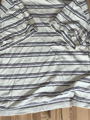 Randig vit t-shirt från arket  - Vit t-shirt från arket med tunna svarta och lila horisontella ränder. Klassisk rund halsringning och korta ärmar. Tillverkad i mjuk bomull som känns skön mot huden. Perfekt för en avslappnad och stilren look.