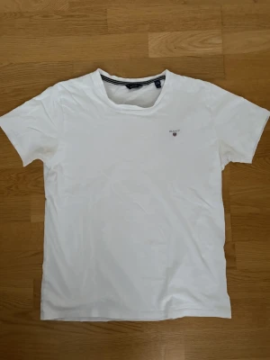 Vit t-shirt från Gant - Klassisk vit t-shirt från Gant med liten logga på bröstet. T-shirten har rund halsringning och korta ärmar. Perfekt basic-plagg i mjuk bomull som passar till allt. Storleken motsvarar S i herr. Smått missfärgad under armarna pga solkräm som man ser i bilden, lätt att ta bort själv. Tveka inte på att höra av dig vid frågor eller funderingar kring passform mm. 