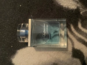 Blå parfym från Baldessarini - Säljer en snygg parfym från Baldessarini i en stilren, rektangulär glasflaska med blått innehåll och genomskinligt lock. Flaskan har silverfärgade detaljer och en modern design som känns lyxig och fräsch.