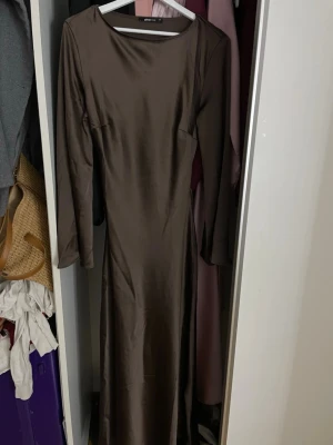 Brun långärmad långklänning - Snygg brun långklänning med lång ärm och rund halsringning. Klänningen har en elegant, enkel design och är tillverkad i ett glansigt material som ger en lyxig känsla. Perfekt för dig som gillar stilrena plagg med en modern touch.