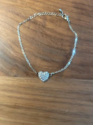 Silverfärgat armband med hjärta - Snyggt armband i silverfärg med en gnistrande hjärtberlock täckt av små pärlor. Kedjan är tunn och smidig med ett klassiskt lås. Perfekt accessoar för dig som gillar stilrena detaljer och lite extra bling.