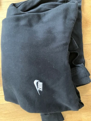 Svarta mjukisbyxor från Nike - Svarta mjukisbyxor från Nike med klassisk broderad logga framtill. Byxorna har en avslappnad passform och är tillverkade i mjukt material som känns skönt mot huden. Perfekta för chill eller träning. Elastisk midja och dragsko för bästa komfort.