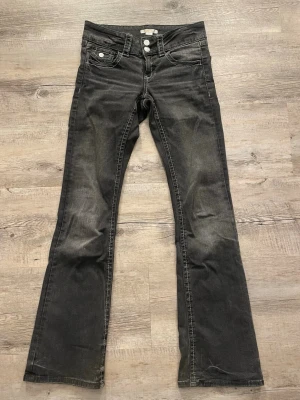 Svarta bootcut jeans med dubbla knappar - Säljer ett par svarta bootcut jeans med dubbla knappar i midjan och vita kontrastsömmar. Jeansen har klassiska fem fickor och lätt slitna detaljer för en cool look. Dem har gått upp lite på bakfickan