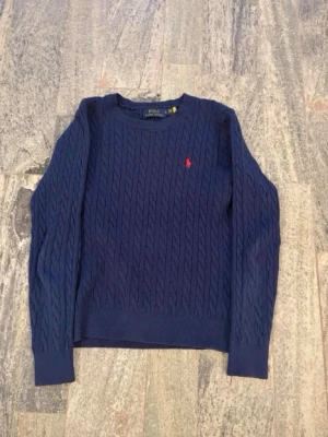 Mörkblå kabelstickad tröja Polo Ralph Lauren - Kabelstickad mörkblå tröja från Polo Ralph Lauren med rund halsringning och röd broderad logga på bröstet. Tröjan har långa ärmar och ribbade muddar. Klassisk och stilren design i mjukt material.