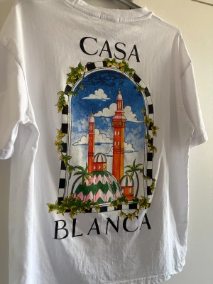 Vit t-shirt från Casablanca - Vit t-shirt från Casablanca med färgglad grafisk print på både bröstet och ryggen. Motivet visar två torn och palmer inramade av en båge med blad. Klassisk passform och rund hals. Perfekt för dig som gillar statement-plagg och snygga prints.