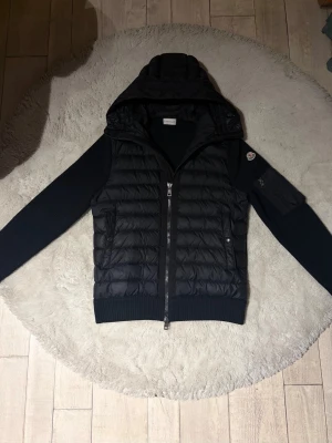 Svart Moncler Cardigan - Svart dunjacka från Moncler med quiltad kropp, stickade ärmar och huva. Jackan har dragkedja framtill, ribbade muddar och en ikonisk Moncler-logga på ärmen, osynlig flaw. Perfekt för dig som gillar en stilren look. True to size (passar M), självklart äkta, priset är inte låst. Hör av vid intresse😄