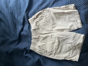 Beige linneshorts med resårmidja - Säljer ett par beige shorts i linne med avslappnad passform och resår i midjan. Shortsen har fickor både fram och bak, perfekta för chill dagar. Snyggt basic-plagg som funkar till det mesta!