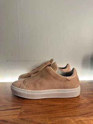 Rosa Axel Arigato Skor - Stilrena rosa sneakers från Axel Arigato i mjuk mocka med vit sula. Minimalistisk design utan synliga snören och diskret logga på sidan. Perfekta för dig som gillar en clean och modern look.