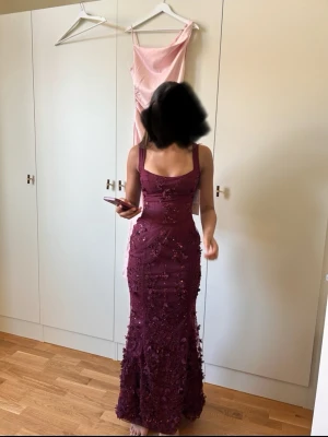 Vinröd långklänning med snörning - Säljer en vinröd långklänning med smala axelband, snörning i ryggen och dekorerad med blommiga applikationer och paljetter. Klänningen har figurnära passform och v-ringad urringning framtill. Perfekt för bal eller festliga tillfällen.