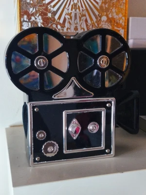🎥 CC-inspired Movie Camera Clutch 🖤✨ - Such a fun and eye-catching statement piece! Designed in the shape of a vintage movie camera with glossy black finish, silver-tone details and a detachable chain strap. It works perfectly as both a clutch and a shoulder bag for parties, dinners, events or whenever you want something a little different.  The bag fits your phone and smaller everyday essentials such as keys, cards and lipstick. Magnetic closure and lined interior.