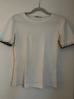 Vit t-shirt med spetsdetalj - Snygg vit t-shirt med korta ärmar och unik svart spetsdetalj längst ut på ärmsluten. T-shirten har en klassisk rund halsringning och är gjord i mjuk bomull som känns skön mot huden. Perfekt för dig som gillar stilrena plagg med en twist.