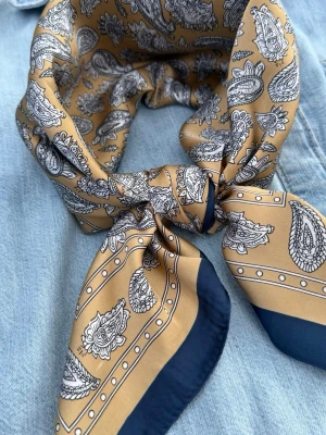 Elegant scarf i camel & marinblått paisley – satinfeeling 70x70 cm - Exklusiv scarf i sofistikerade toner av varm camel, djup marinblå och ivory med detaljrikt paisleymönster och elegant kontrasterande kant. Den silkeslika satinfeelingen ger ett vackert fall och en lyxig lyster som omedelbart lyfter en outfit.  En tidlös accessoar med exklusiv känsla – lika självklar till en krispig skjorta och kavaj som till stickat, trenchcoat eller en enkel sommarlook. Färgkombinationen ger en diskret couturekänsla med inspiration från klassiskt europeiskt mode och vintage sil