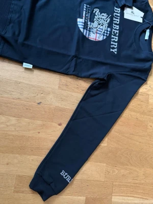Svart sweatshirt från Burberry - Svart sweatshirt från Burberry med broderad logga och rutig detalj framtill. Tröjan har rund halsringning, långa ärmar och ribbade muddar. Klassisk och stilren design med Burberry-tryck även på ärmen.