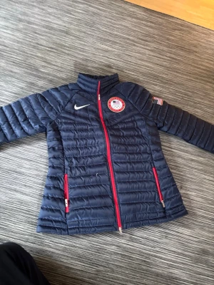 Blå Nike pufferjacka Team USA - Mörkblå pufferjacka från Nike med Team USA-märke på bröstet och amerikansk flagga på ärmen. Jackan har röda dragkedjor och två fickor framtill. Perfekt för kalla vinterdagar och har en sportig look. Pris kan självklart diskuteras 