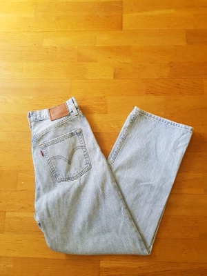 Ljusblå Levi's raka jeans - Säljer ett par klassiska ljusblå jeans från Levi's med rak passform och hög midja. Jeansen har fem fickor, Levi's ikoniska läderpatch bak och röd etikett på bakfickan. Tillverkade i 100% bomull för en autentisk denimkänsla.
