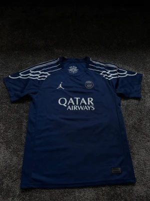 PSG x Jordan blå matchtröja - Mörkblå PSG matchtröja i samarbete med Jordan, med vita detaljer och vingmönster på axlarna. Tryck med Qatar Airways på bröstet och PSG-logga samt Jumpman-logga. Perfekt för fotbollsträning eller att bära som supporter.