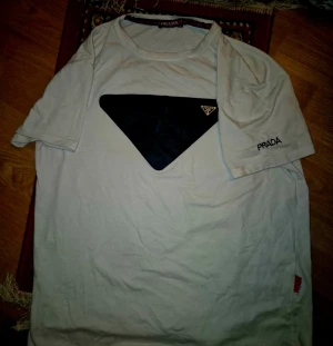 Vit t-shirt från Prada med svart detalj - Vit t-shirt från Prada med en stor svart triangel på bröstet och diskret Prada-logga på ärmen. Klassisk rund hals och korta ärmar. Perfekt för dig som gillar stilrena plagg med en twist. Xl men sitter mer som en m konstiga storlekar... värderad till 800-1500