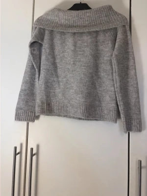 Grå stickad tröja från Gina Tricot - Mysig grå stickad tröja med bred, ribbstickad krage som kan vikas ner över axlarna. Tröjan har långa ärmar och ribbade muddar vid ärmslut och nederkant. Perfekt för kyliga dagar och enkel att matcha med jeans eller kjol.