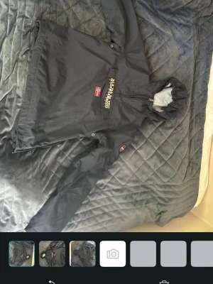 Svart vindjacka från Napapijri - Snygg svart vindjacka från Napapijri med halv dragkedja, stor magficka och norsk flagga på bröstet. Jackan har huva med dragsko och är tillverkad i ett vattenavvisande material, perfekt för blåsiga dagar. MEN SPECIELT I SKIDBACKEN