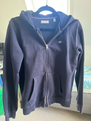 Mörkblå hoodie med dragkedja från Gant - Mörkblå hoodie från Gant med dragkedja framtill, två fickor och broderad logga på bröstet. Klassisk modell med huva och ribbade muddar. 