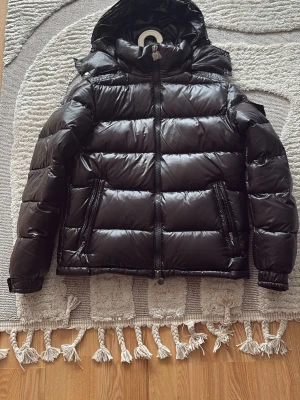 Svart Moncler Maya - Snygg svart pufferjacka med glansig finish och huva. Jackan har två stora dragkedjefickor framtill och breda kanaler för extra värme. Perfekt för kalla dagar och har en modern, boxig passform. Materialet är troligtvis syntet med fyllning för värme.