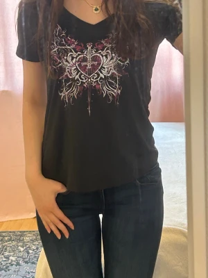 Svart t-shirt med hjärtmotiv och svärd - Cool svart t-shirt med v-ringning och tryck av ett hjärta genomborrat av ett svärd, omgiven av vita och rosa detaljer. 🖤
