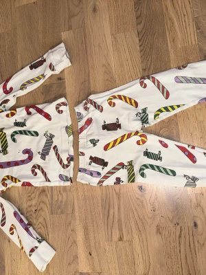 Vit pyjamas med polkagrisar - Säljer en vit pyjamas med färgglada polkagrisar och karameller i rött, grönt, gult, lila och brunt. Setet består av långärmad tröja och byxor med resår i midjan. Perfekt för mysiga kvällar och julkänsla.