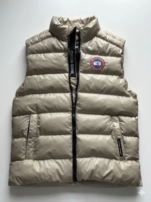Beige dunväst från Canada Goose - Snygg beige dunväst från Canada Goose med klassisk logga på bröstet och svart dragkedja med märkets namn. Västen har två fickor framtill och är tillverkad i ett glansigt, quiltat material som håller värmen. Perfekt för lager-på-lager-stil. Skick 8/10 finns lite färg på den. Storlek s 