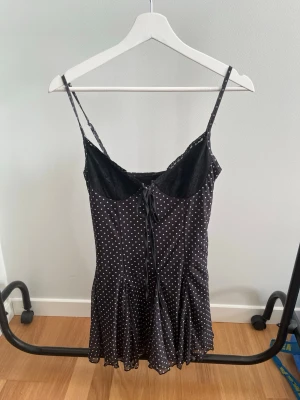 Svart prickig kort klänning med snörning - Svart kort klänning med vita prickar och smala axelband. Klänningen har snörning framtill och spetsdetaljer vid bysten. Lätt utsvängd nederdel ger en feminin siluett. Perfekt för dig som gillar en söt och trendig stil.