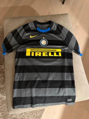 Inter Milan bortatröja Nike svart/grå - Inter Milan bortatröja från Nike med breda svarta och gråa ränder, gula detaljer och Pirelli-logga på bröstet. Tröjan har klubbmärke, rund hals med blå inslag och är tillverkad i lätt funktionsmaterial. Perfekt för fotbollsträning eller match.