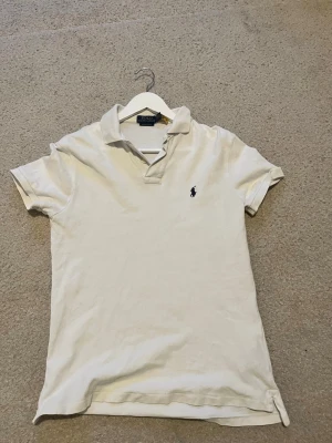 Vit pikétröja från Ralph Lauren - Klassisk vit pikétröja från Ralph Lauren med korta ärmar och den ikoniska loggan broderad på bröstet. Tröjan har krage och knappar vid halsen. Perfekt till jeans eller shorts för en clean och stilren look.