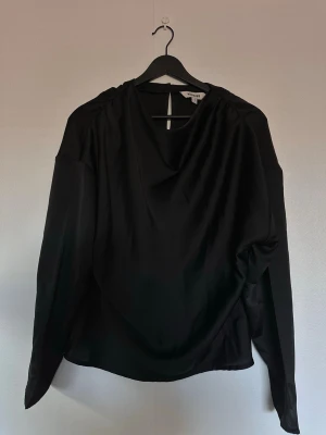 Svart satinsblus från Kappahl  - Stilren svart blus i satin från Kappahl med draperad halsringning och långa ärmar. Blusen har en lätt glansig finish och en avslappnad passform, perfekt att styla med jeans eller kjol. Materialet känns mjukt och följsamt mot huden.