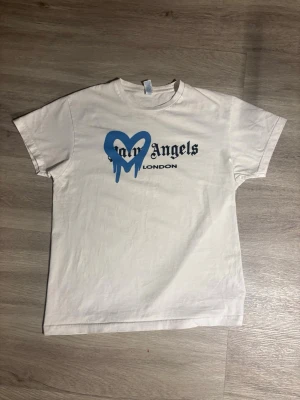 Vit Palm Angels t-shirt med blått hjärta - Snygg vit t-shirt från Palm Angels med tryck på bröstet. Framsidan har ett blått, droppande hjärta och svart text med 'Palm Angels London'. Klassisk passform och rund hals. Perfekt för en trendig streetwear-look.