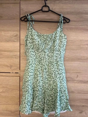 Grön blommig kortklänning med shorts  - Söt ljusgrön kortklänning med blommigt mönster och tunna axelband som knyts upptill. Klänningen har shorts under med en lätt volang i nederkanten och smickrande passform. Perfekt för varma dagar och sommarens alla tillfällen.