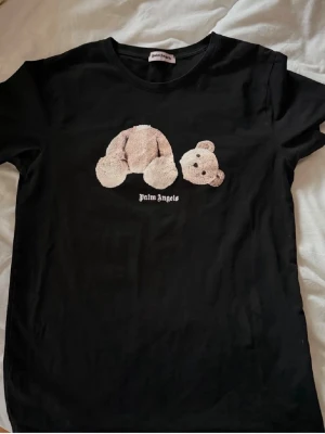 Svart Palm Angels t-shirt med nalle - Svart t-shirt från Palm Angels med ett broderat motiv av en nallebjörn på bröstet och logotyp under. Klassisk rund hals och korta ärmar. Perfekt för dig som gillar streetwear och vill sticka ut med en unik detalj.