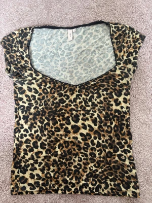Leopardmönstrad topp med v-ringning - Snygg topp med leopardmönster och  v-ringning. Har korta ärmar och är gjord i ett mjukt, stretchigt material. Svart spetskant runt halsen. Från Nelly i storlek S.💗