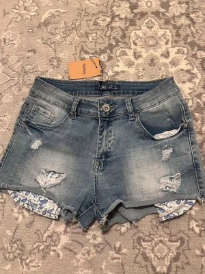 Blå jeansshorts från Toxik3 - Snygga blå jeansshorts från Toxik3 med slitna detaljer och råa kanter. Shortsen har klassisk femficksdesign och mönstrat tyg som sticker fram vid bensluten. Köpta på Sellpy men aldrig använda av mig!💝Storlek 40 men dom passar mig som vanligtvis bär 36-38. Har också stretch. 