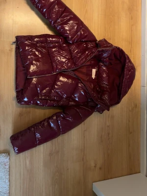 Nelly vinröd glansig pufferjacka med huva - Säljer en vinröd pufferjacka med glansig finish och huva ifrån Nelly. Knappt använd i mycket bra skick.
