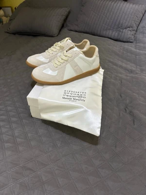 Maison Margiela vita sneakers - Maison Margiela sneakers i vitt och beige med klassisk låg profil och gummisula. Ovandel i läder och mocka, med vita och beige detaljer samt matchande snörning. Perfekt för dig som gillar stilrena och exklusiva sneakers.