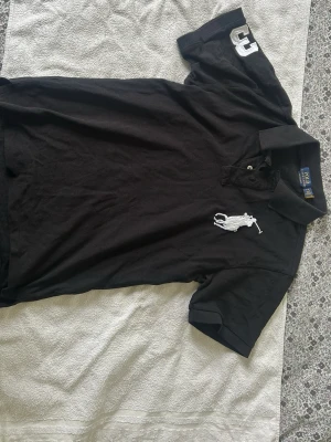 Svart pikétröja från Ralph Lauren - Svart pikétröja från Ralph Lauren med stor vit broderad logga på bröstet och siffran 3 på ärmen. Klassisk krage och knäppning framtill. Perfekt för en sportig och stilren look.