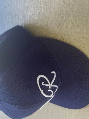 Mörkblå Vonroe keps - Säljer en mörkblå keps från Vonroe med vit broderad logga framtill och detaljer på sidan. Klassisk snapback-modell med rak skärm och ventilerande hål. Snygg streetwear-vibe och passar perfekt till en avslappnad stil.