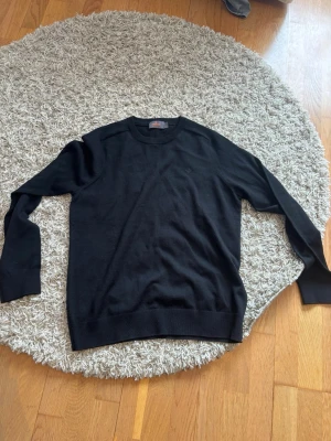 Svart Morris tröja M - Svart stickad tröja med rund halsringning och diskret broderad logga på bröstet. Tröjan har långa ärmar och ribbade muddar vid ärmslut och nederkant. Perfekt för en stilren och enkel look. Tveka inte att höra av dig vid frågor eller pris. 